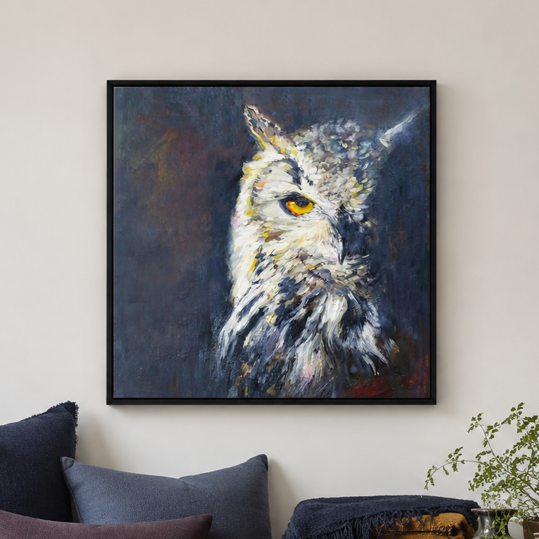 A framed square canvas painting of an owl's head in profile, displayed on a light beige wall above a sofa with dark blue and gray cushions. The painting features textured brushstrokes in white, gray, black, and subtle warm tones, with a yellow eye.