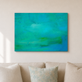 An abstract canvas wall art print featuring shades of teal, turquoise, and green, displayed on a light beige wall above a neutral-toned sofa with decorative pillows.