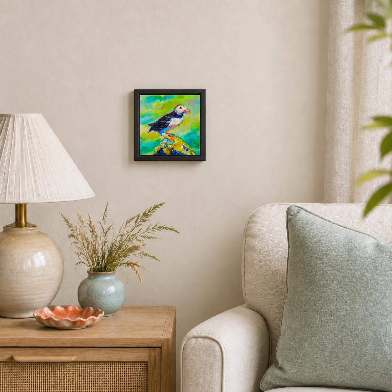 A small framed painting of a puffin hanging on a beige wall in a styled living room corner with a lamp, vase, side table, and part of a sofa visible.