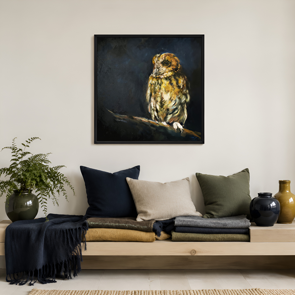 A framed square canvas painting of an owl perched on a branch, displayed on a light beige wall above a wooden bench with pillows and folded textiles in a styled interior setting.