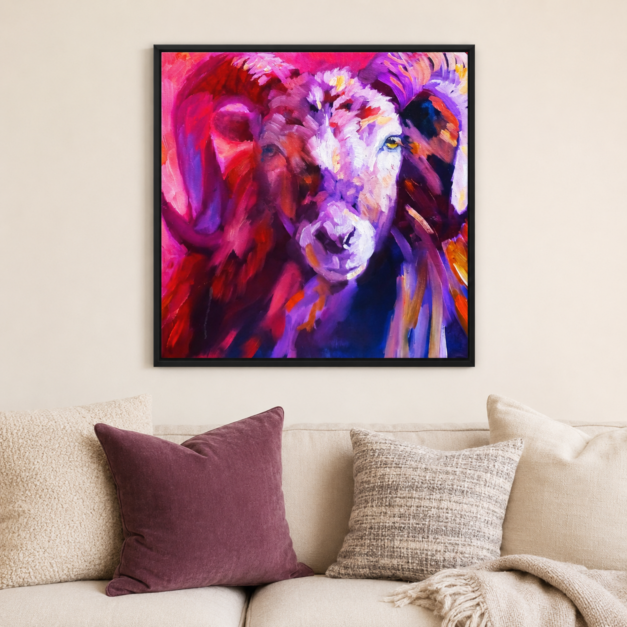 A colorful framed wall art print depicting a stylized ram portrait in vivid pink, purple, red, and blue tones, hanging above a beige sofa with decorative pillows in a living room setting.
