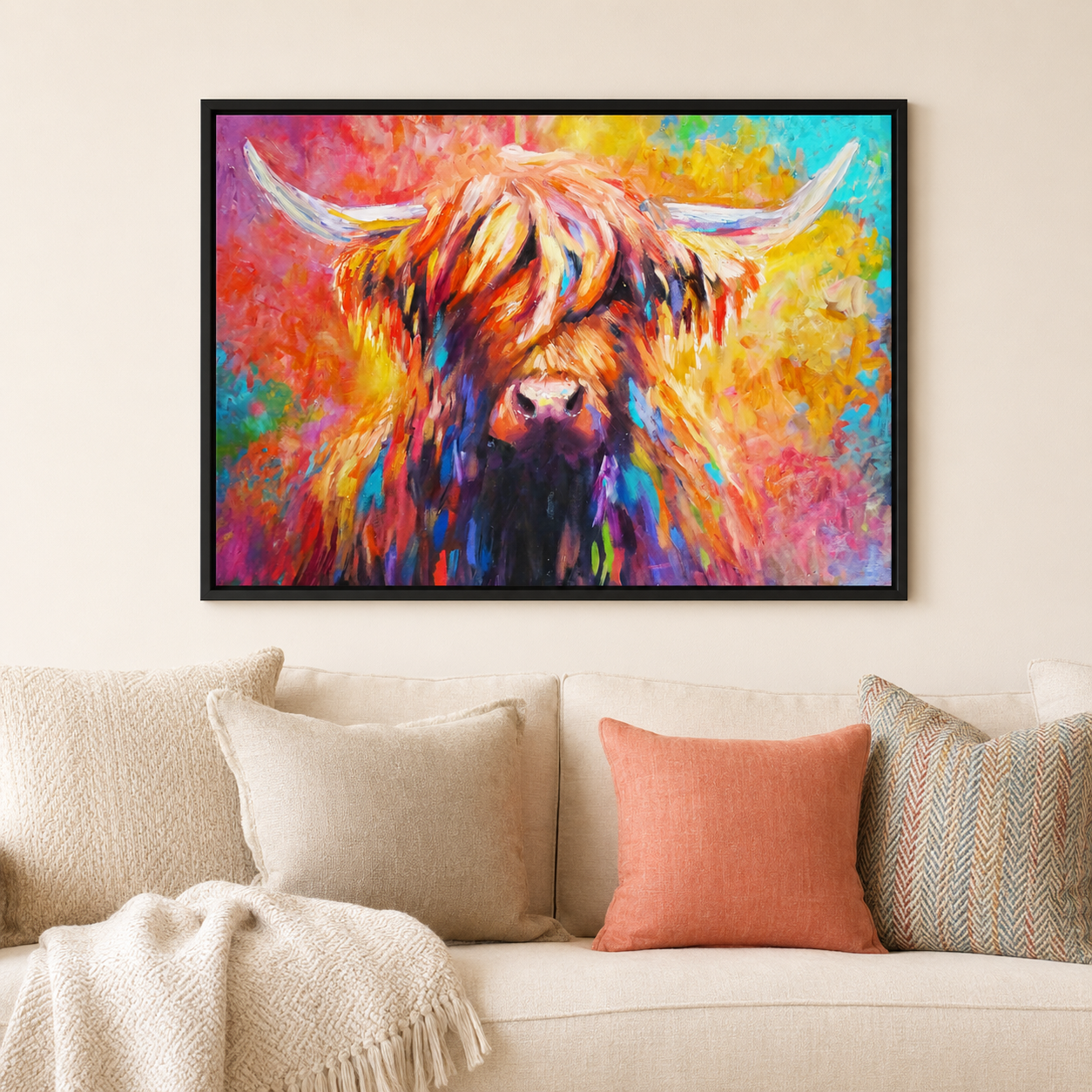 A colorful framed wall art print of a Highland cow hangs above a beige sofa with cream and coral pillows in a living room setting.