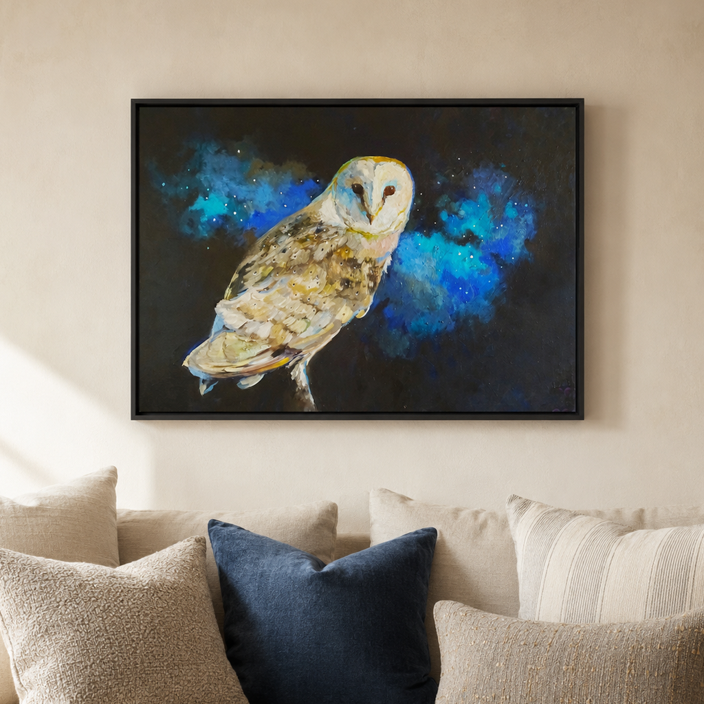 A framed wall art print showing a barn owl against a dark blue and black cosmic background, displayed above a beige sofa with decorative pillows in a living room setting.