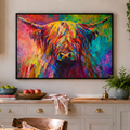A colorful framed canvas print of a Highland cow hangs on a beige wall in a bright, modern kitchen. The artwork features a vibrant, textured paint style in a rainbow of colors including yellow, orange, red, pink, purple, and blue.