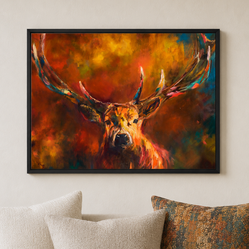 A framed wall art print showing a stylized deer with large antlers in warm fiery tones, displayed above a sofa with cream and patterned pillows in a living room setting.