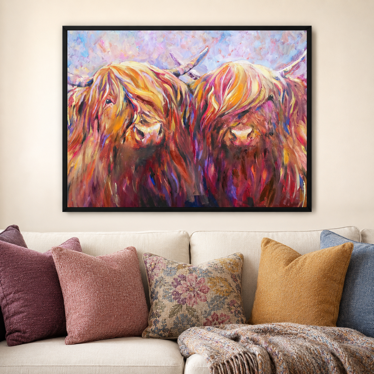 A colorful framed painting of two Highland cows hangs on a beige wall above a white sofa with decorative throw pillows and a blanket.