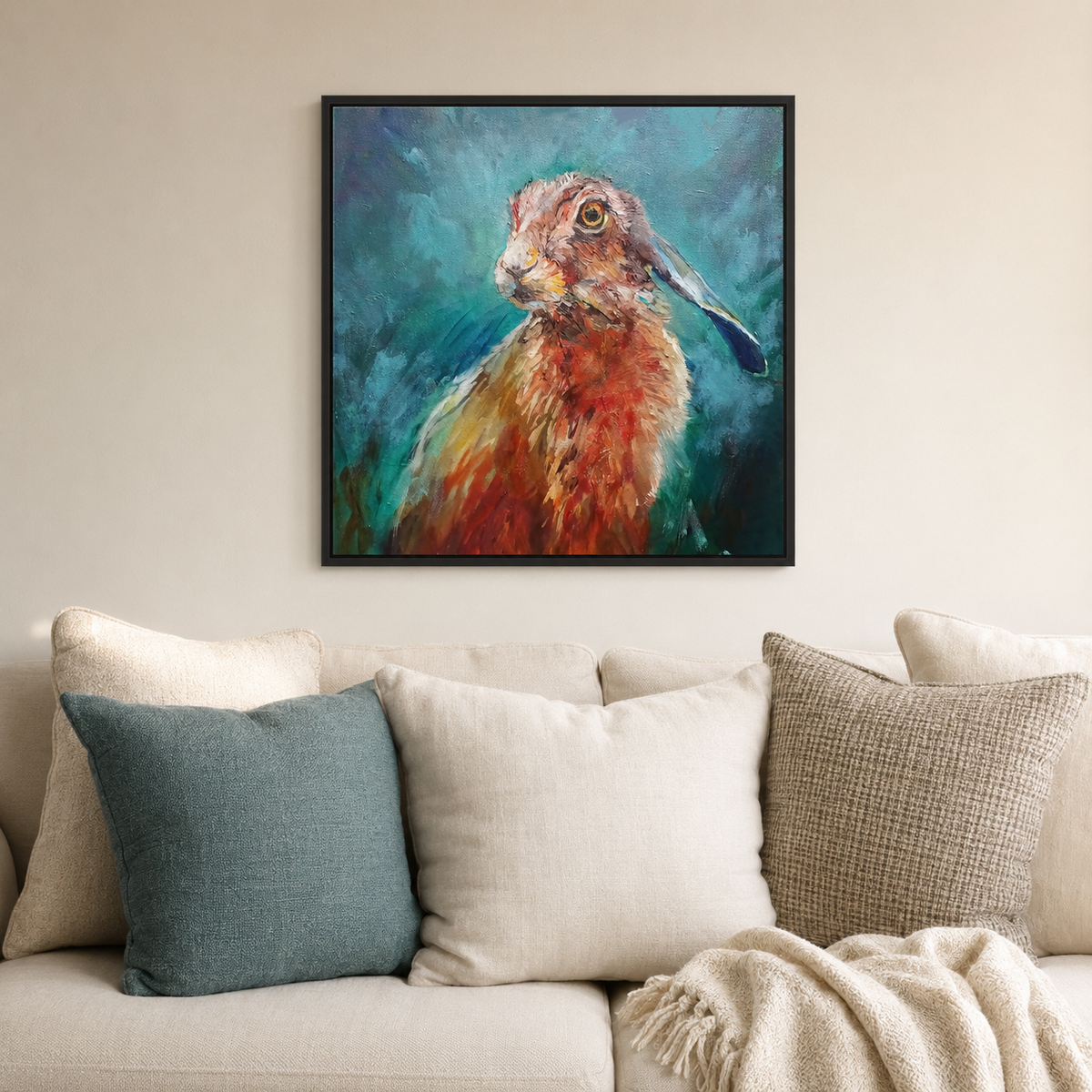 A framed square canvas print of an expressive painted hare in warm orange, brown, and teal tones, hanging on a beige wall above a light sofa with cream and blue pillows.