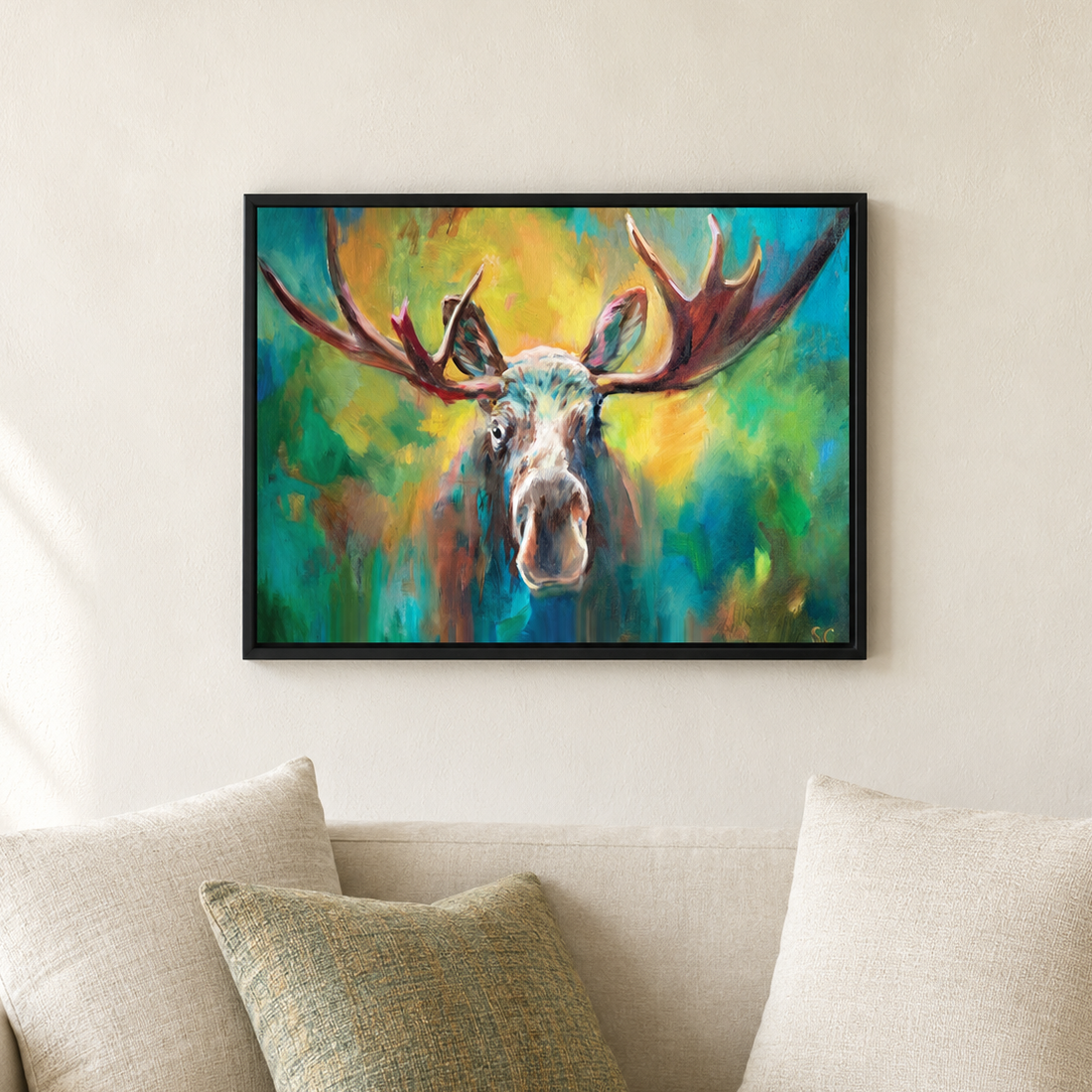 A framed colorful painting of a moose head with large antlers, displayed on a beige wall above a light sofa with pillows.