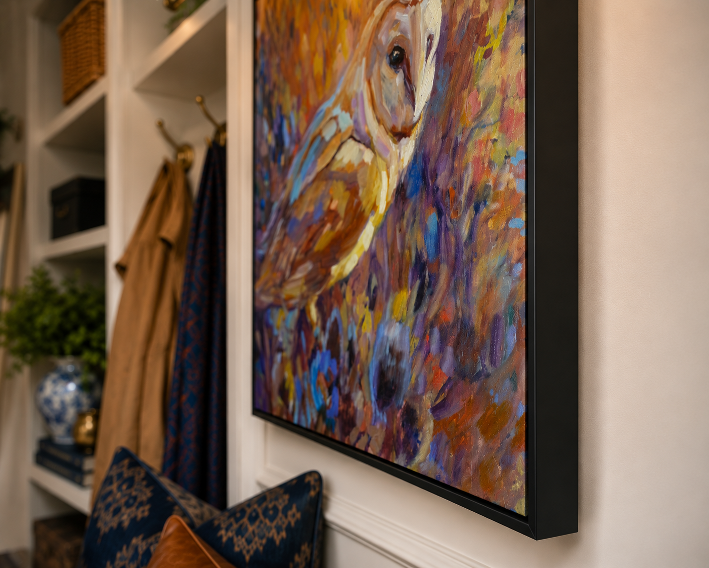 A framed colorful painting of an owl hanging on a white wall in a home entryway, with shelves, a coat rack, and a cushion visible nearby.