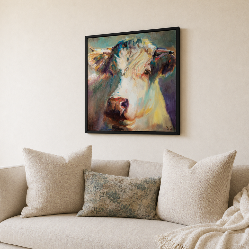 A framed square canvas print of an expressive cow painting hangs on a beige wall above a light beige sofa with cream pillows and a throw blanket.