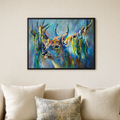 A framed colorful painting of a deer with antlers hanging from its horns, displayed on a beige wall above a sofa with cream and patterned pillows.